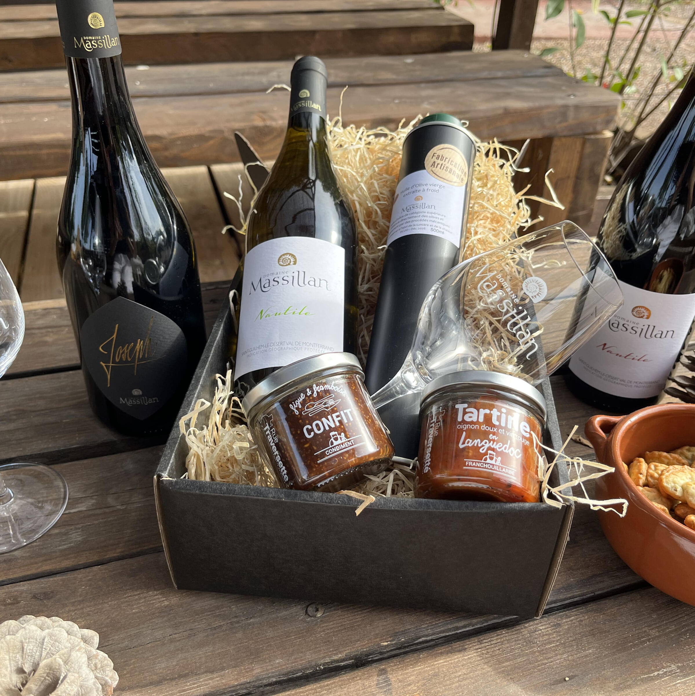 Coffret cadeau du Domaine Massillan composé de vins bio, confit de figue et framboise, tartinade artisanale, huile d’olive vierge et verres gravés, présenté sur une table en bois. Tous nos coffrets sont personnalisables. Photo non contractuelle.